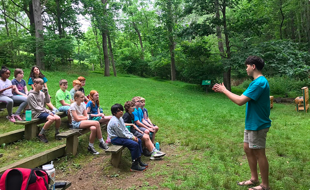 Outdoor educator speaking to a seated group in a wooded area.